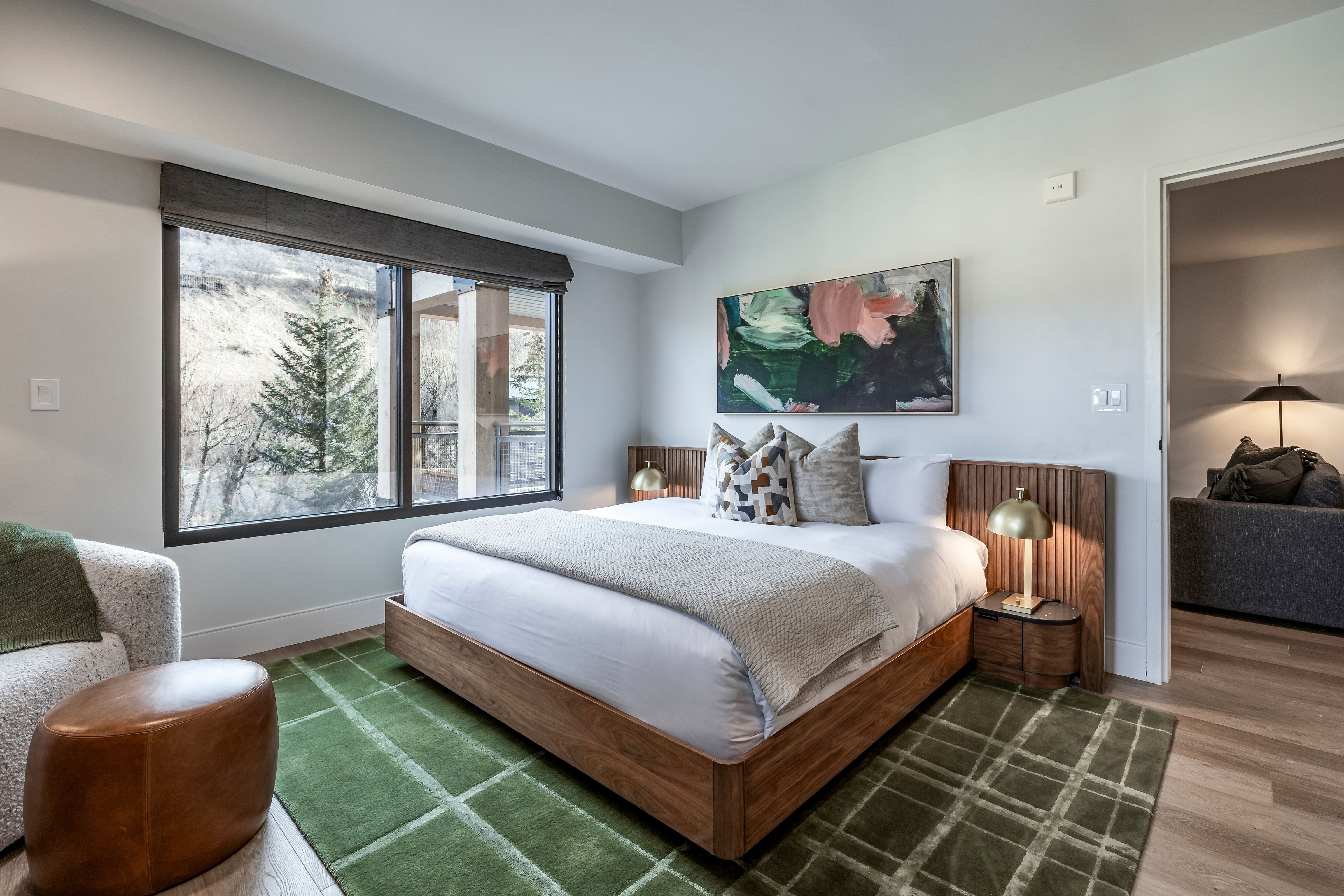 A wide view of a hotel room with a bed, window, artwork, rug, and chair.