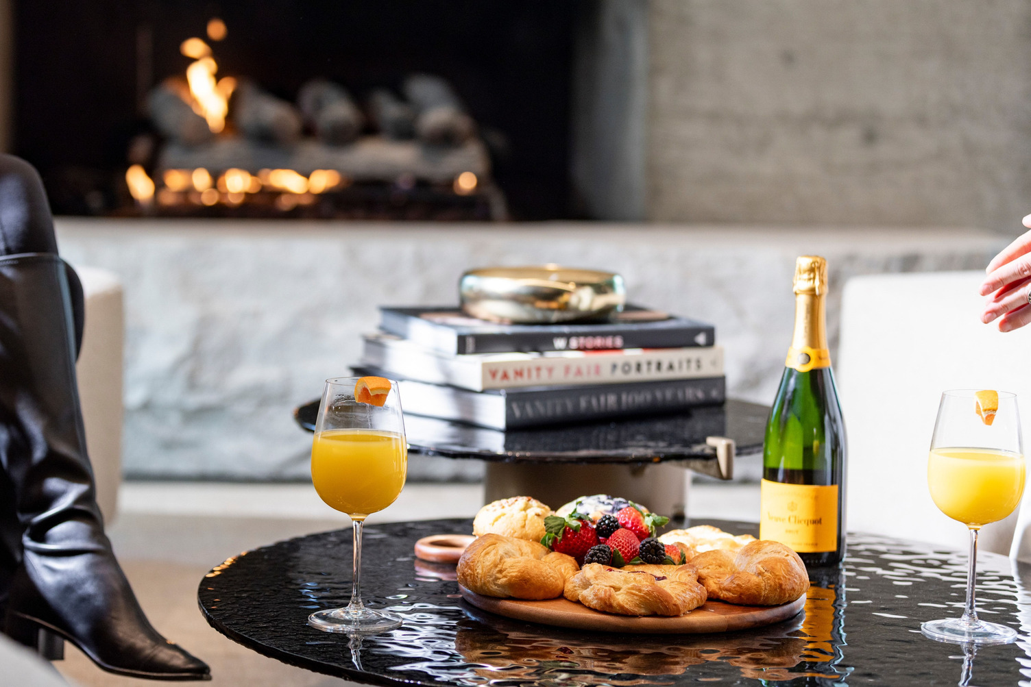 A cozy lobby table set with pastries, wine, and orange juice in front of a fireplace.